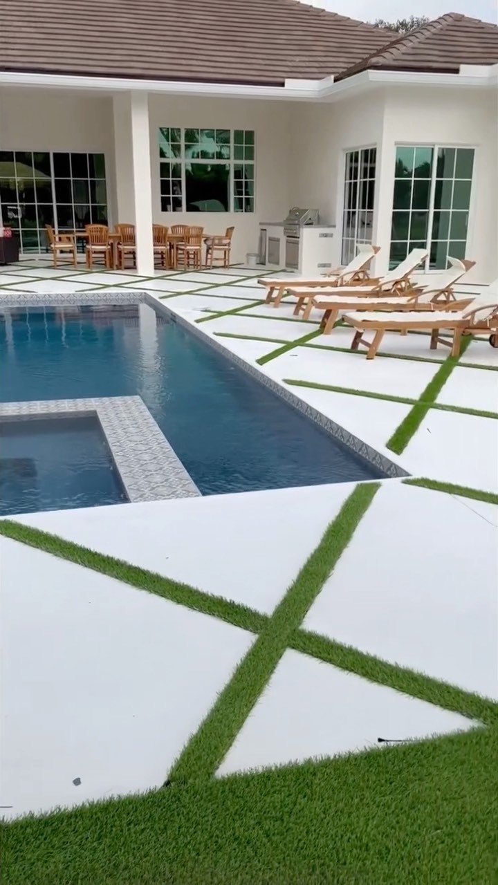 White concrete pool deck with diagonal artificial grass lines, spa, loungers, and outdoor kitchen at a Parkland home